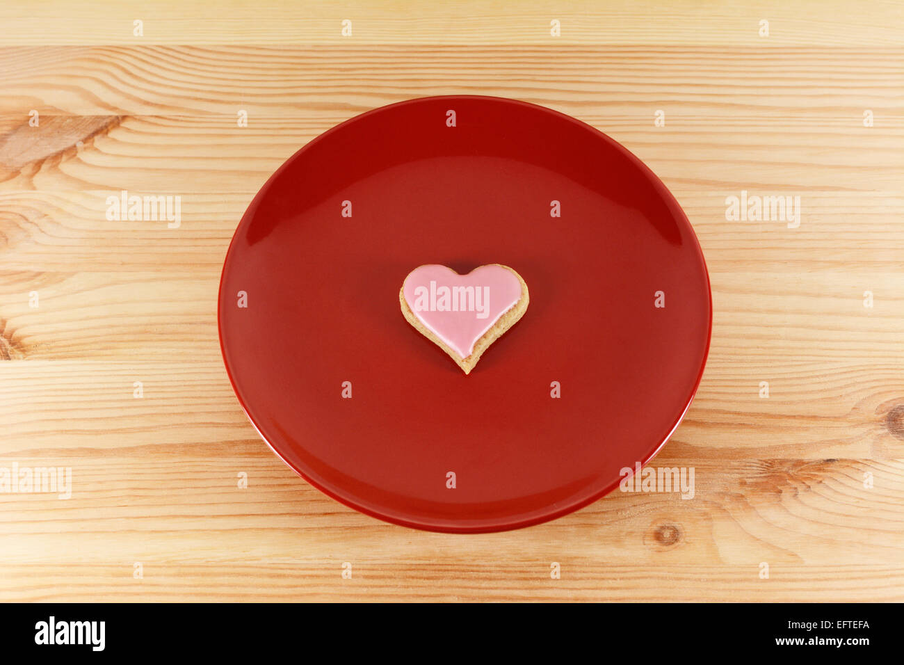 Eine herzförmige Cookie mit rosa Zuckerguss auf einer roten Platte auf einem Holztisch Stockfoto