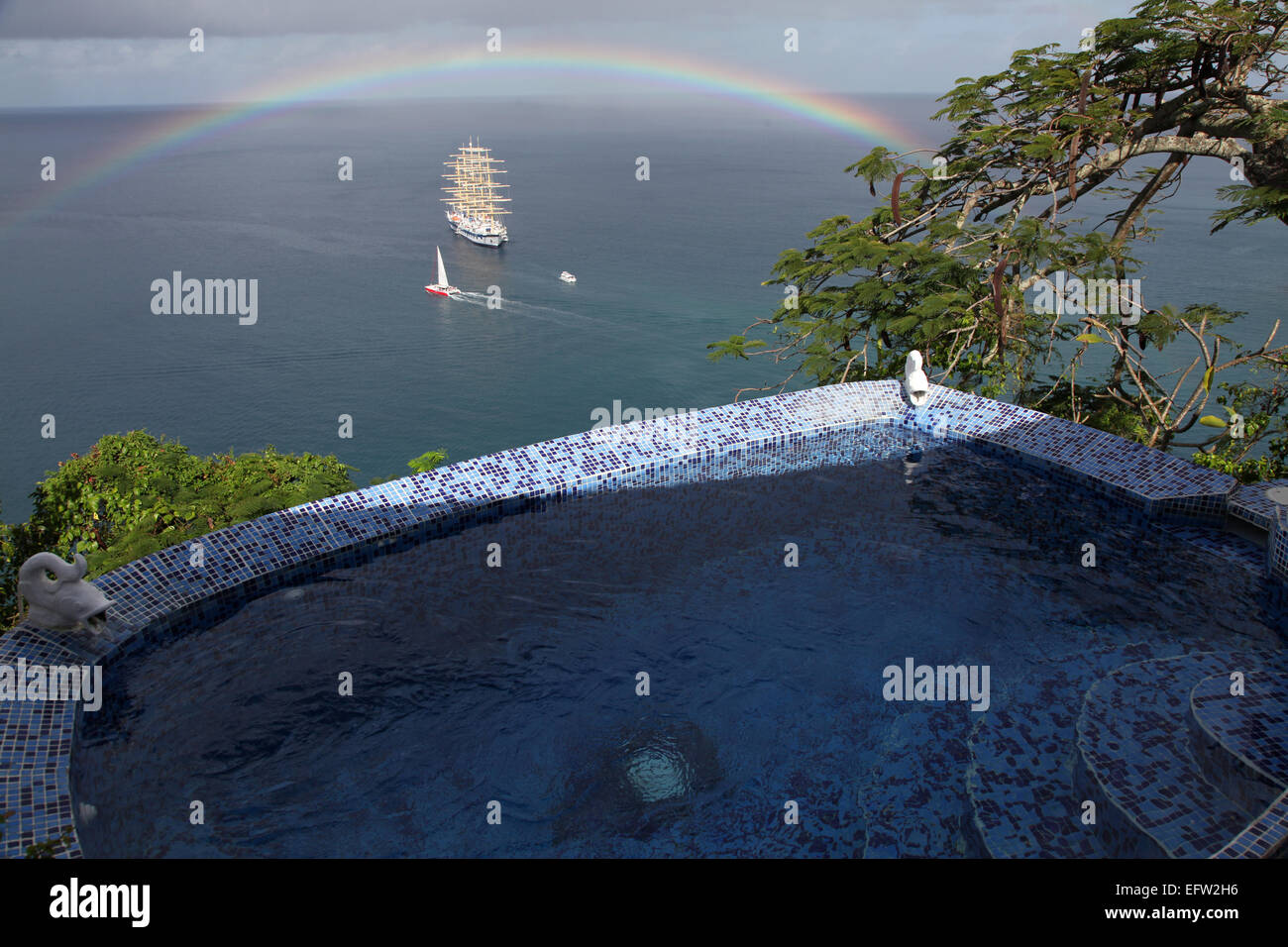 Ein Klipper vertäut aus St. Lucia mit einem Swimmingpool und Johannisbrotbaum im Vordergrund und ein Regenbogen insgesamt. Stockfoto