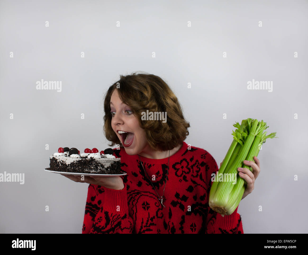 Junge Frau, die versucht zu einer Diät hält, Sellerie in einer Hand und Kuchen in der anderen Stockfoto
