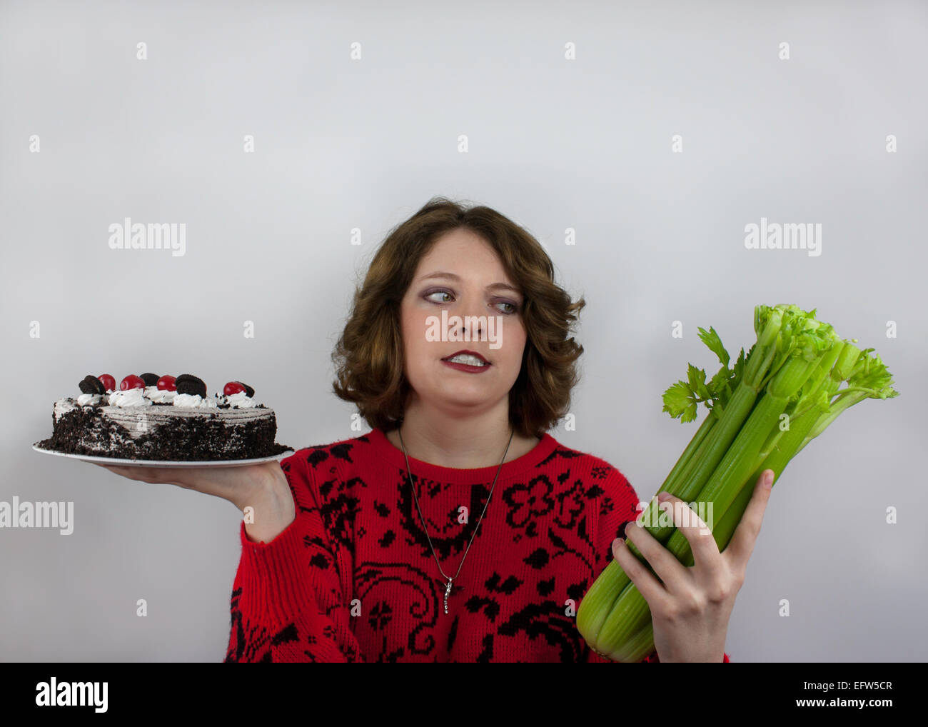 Junge Frau, die versucht zu einer Diät hält, Sellerie in einer Hand und Kuchen in der anderen Stockfoto
