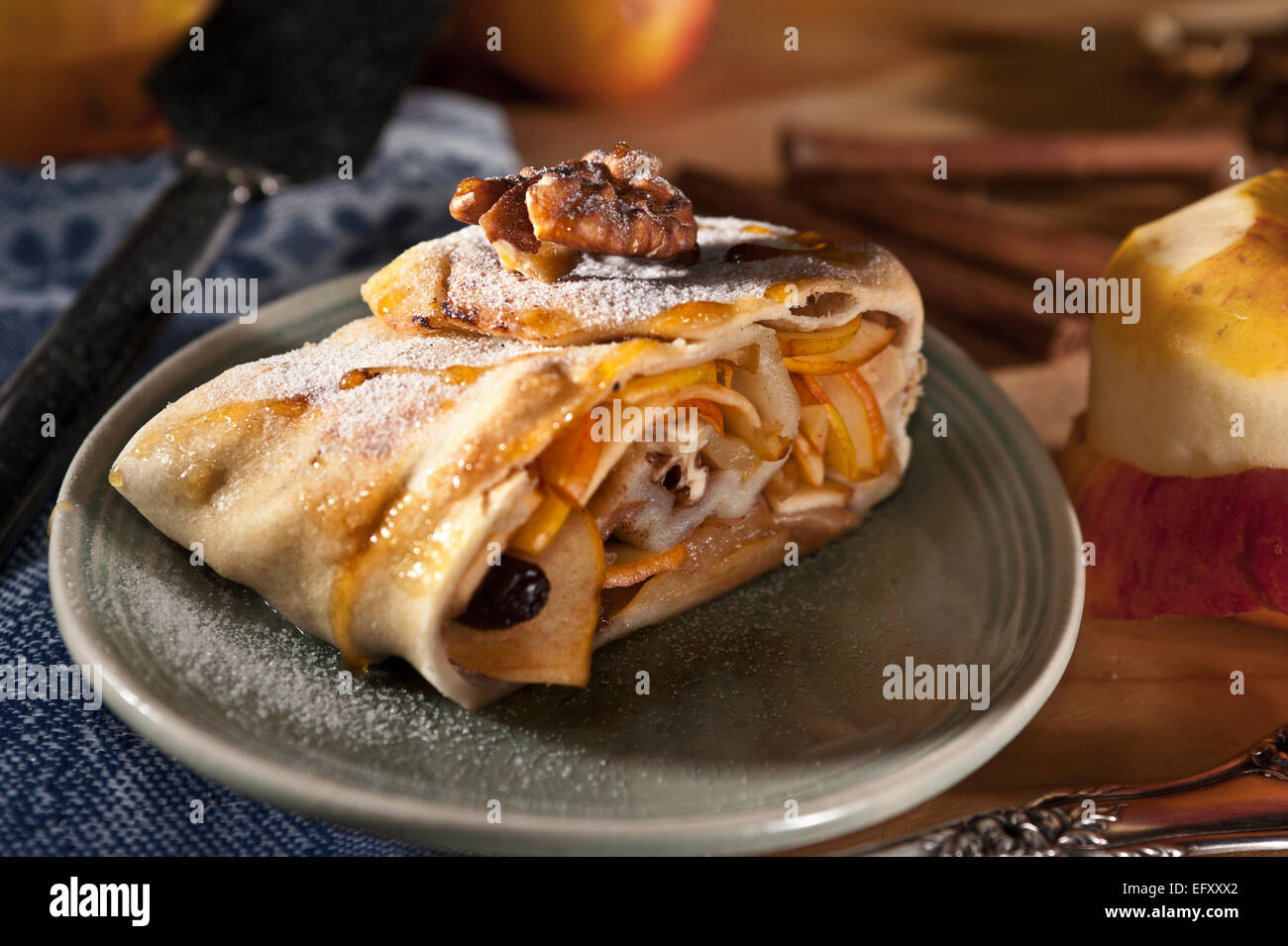 Apfelstrudel mit Walnüssen und Rosinen, serviert auf einem blauen Teller mit Äpfeln und Zimt im Hintergrund. Stockfoto