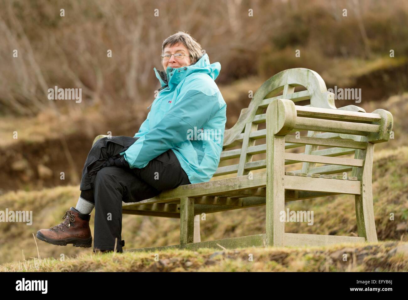 Frau in blauer Jacke sitzt in einer erhöhten große hölzerne Parkbank während eine Sportveranstaltung - Skye V Lovat, Shinty Spiel beobachten. Stockfoto