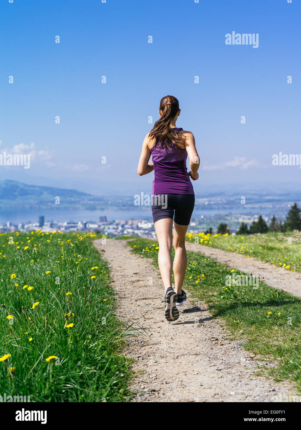 Foto einer jungen Frau, Joggen und trainieren auf einem Feldweg.  See und Stadt in der Ferne. Leichte Bewegungsunschärfe sichtbar. Stockfoto