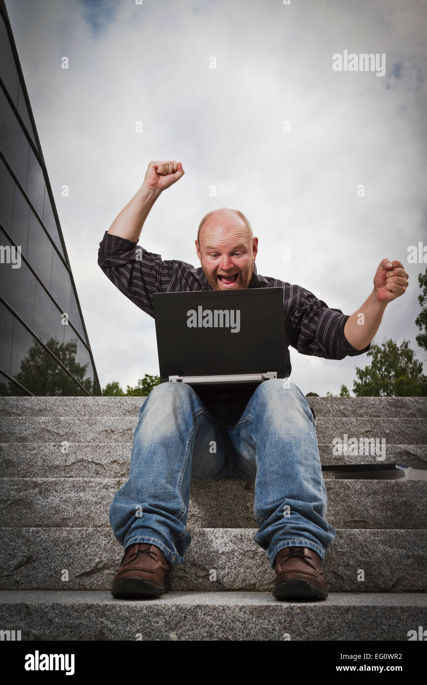 Mann mit seinem Laptop zu feiern eine Leistung oder ein Gewinn. Im Treppen draußen zu sitzen. Stockfoto