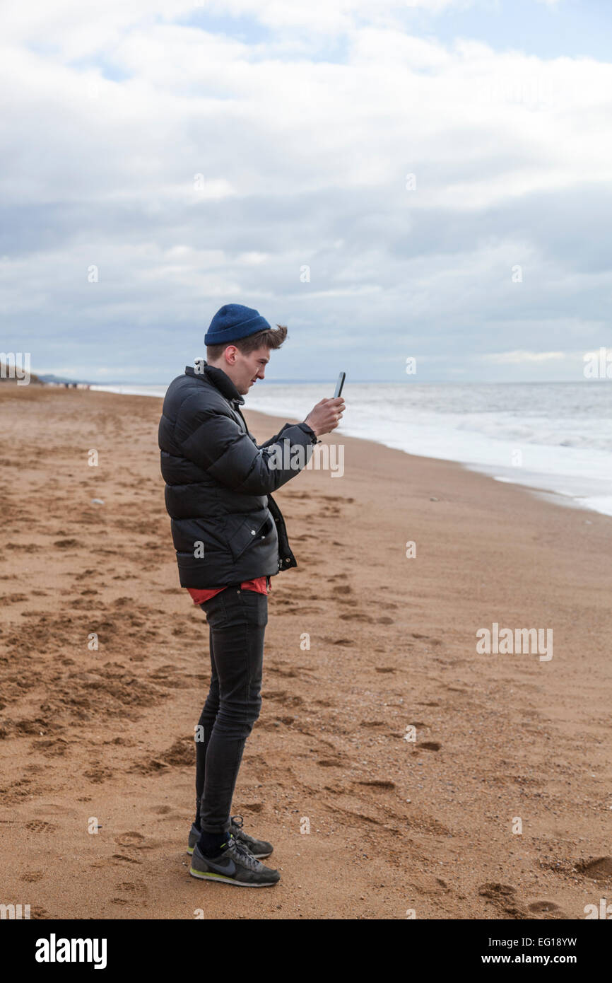 Junger Mann tragen 'Puffa' Jacke und wollene Mütze auf Chesil Beach im Winter mit dem Fotografieren mit einem Ipad. Stockfoto