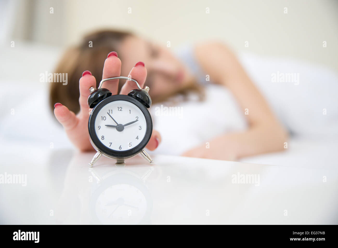 Verschlafene Frau im Bett, die Hand mit Wecker reichen. Uhr im Fokus Stockfoto