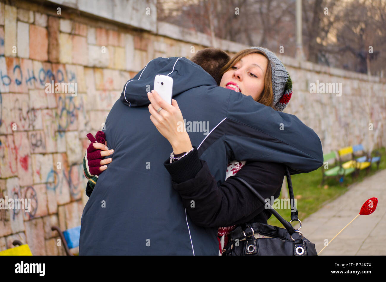 Junge, eine Mädchen umarmt, während sie ihr Smartphone im Freien sieht Stockfoto