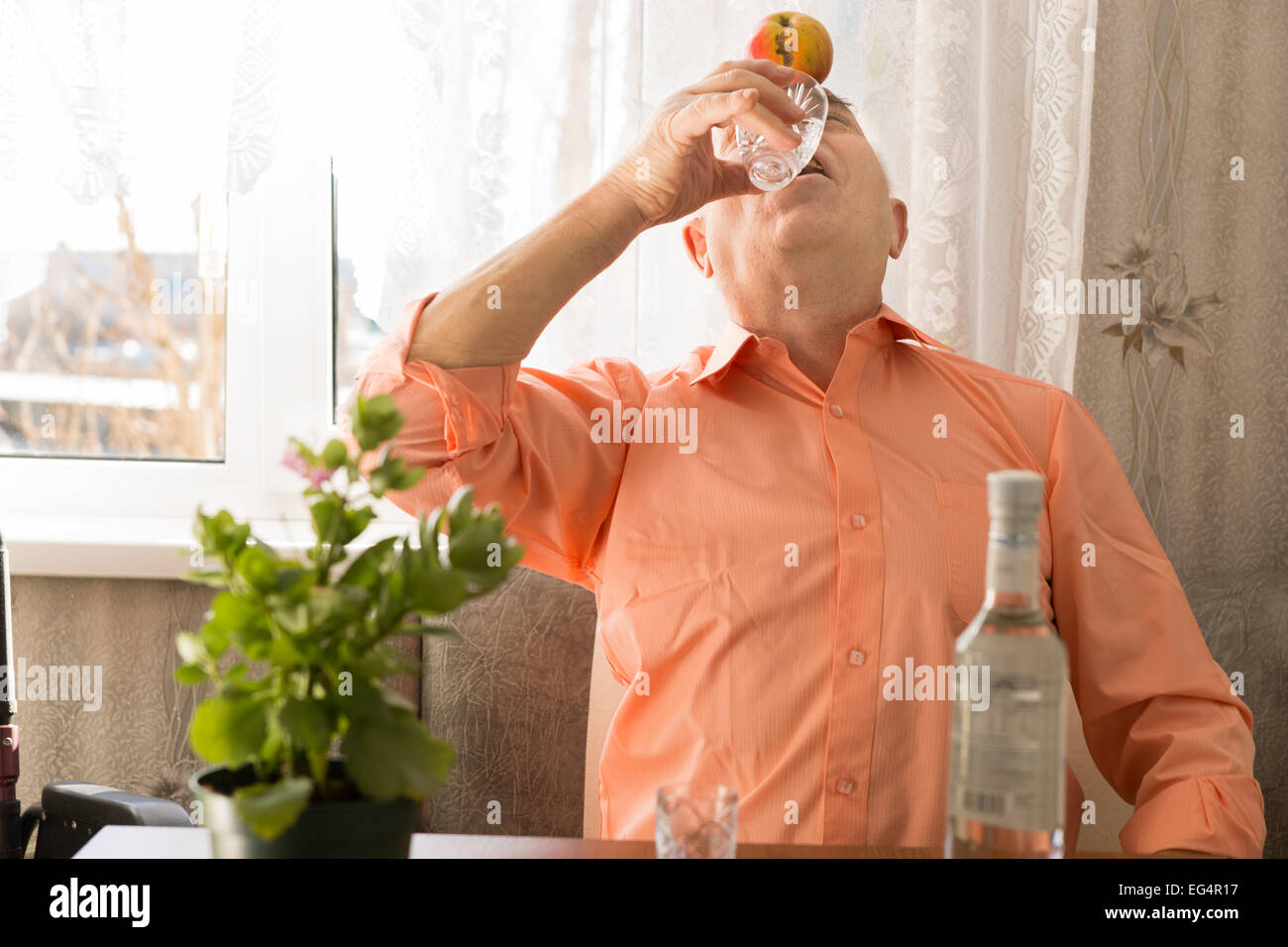 Spielerische ältere Vodka Wein trinken mit Apple auf Stirn, während an der lebenden Bereich nah an das Fenster sitzen hautnah. Stockfoto