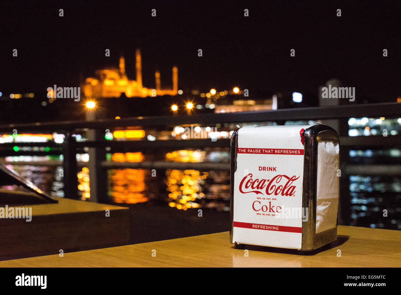 Coca-Cola Servietten Restaurant Galata Bridge Istanbul // ISTANBUL, Türkei – Coca-Cola Servietten in einem Spender auf einem Tisch in einem Restaurant an der Galata Brücke in Istanbul, mit Blick auf das Goldene Horn und mit den Lichtern einer der markanten Moscheen der Stadt im Hintergrund. Stockfoto