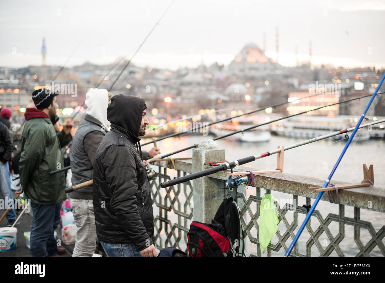 ISTANBUL, Türkei / Türkiye — Fischer säumen den Rand der Galata-Brücke mit ihren Linien über die Seite zum Goldenen Horn. Die Galata-Brücke, die das Goldene Horn überspannt und Eminonu mit Karakoy verbindet, ist eine zweistöckige Brücke, die Straßen-, Straßenbahn- und Fußgängerverkehr auf der obersten Ebene mit Restaurants und Bars auf der darunter liegenden Ebene abwickelt. In der Ferne befindet sich die Suleymaniye-Moschee. Stockfoto