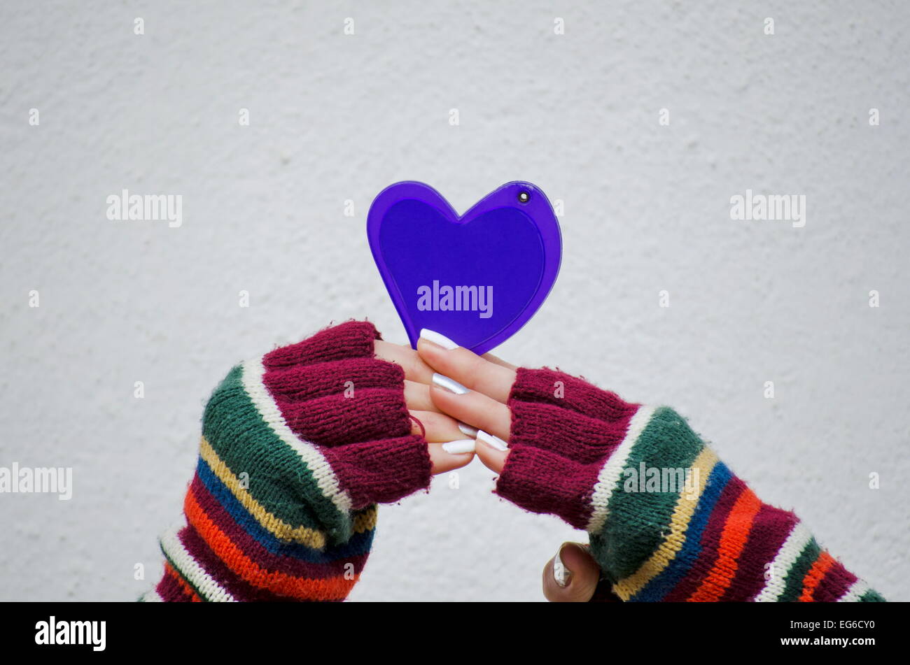 Mädchen in bunte Fäustlinge hält ein lila Herz gegen eine weiße Wand Stockfoto