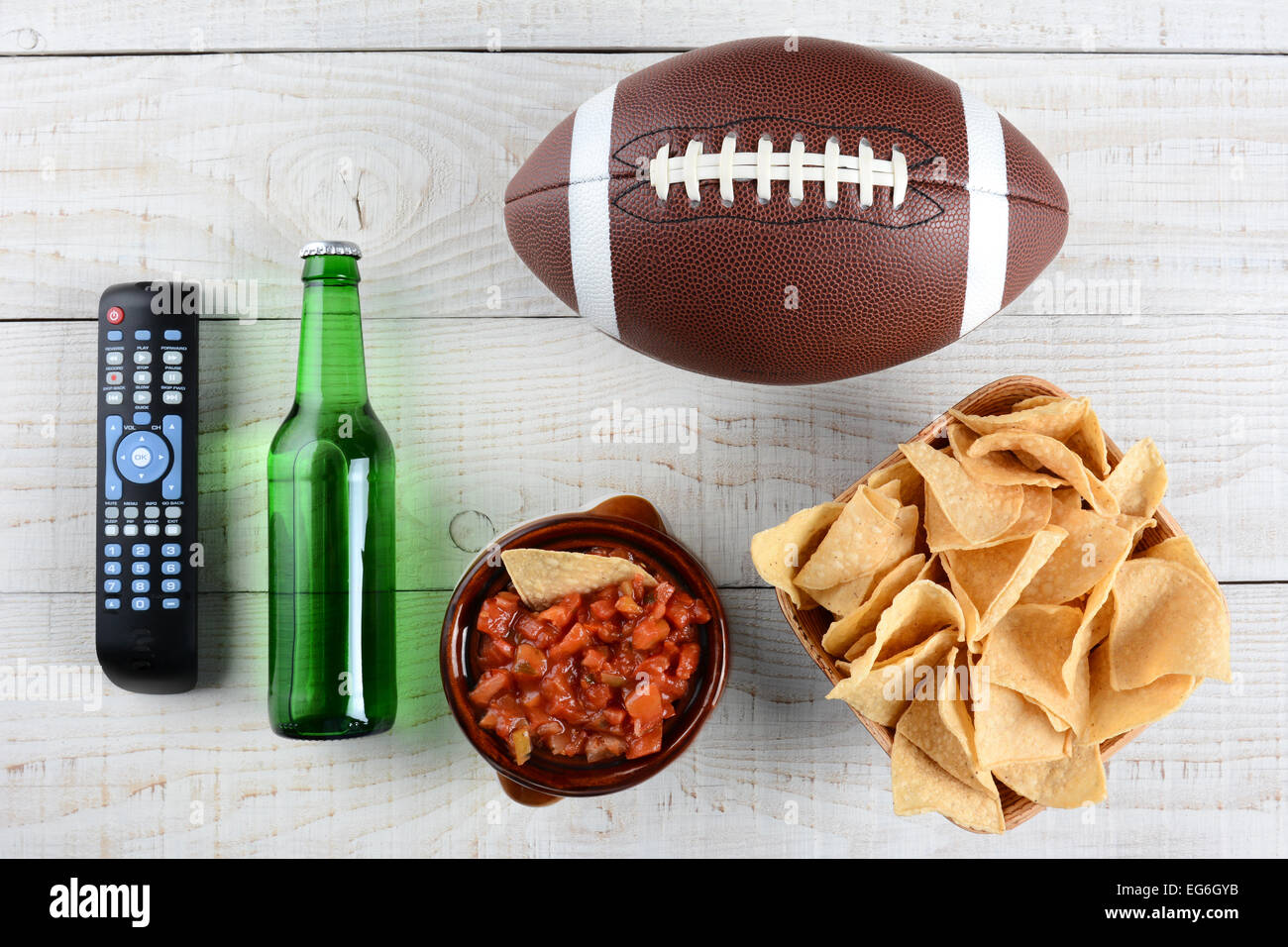 TV-Fernbedienung, Bierflasche, Schüssel mit Chips mit Salsa und ein amerikanisches Fußball auf einem rustikalen weiß getünchten Holzoberfläche. Horizontale Stockfoto