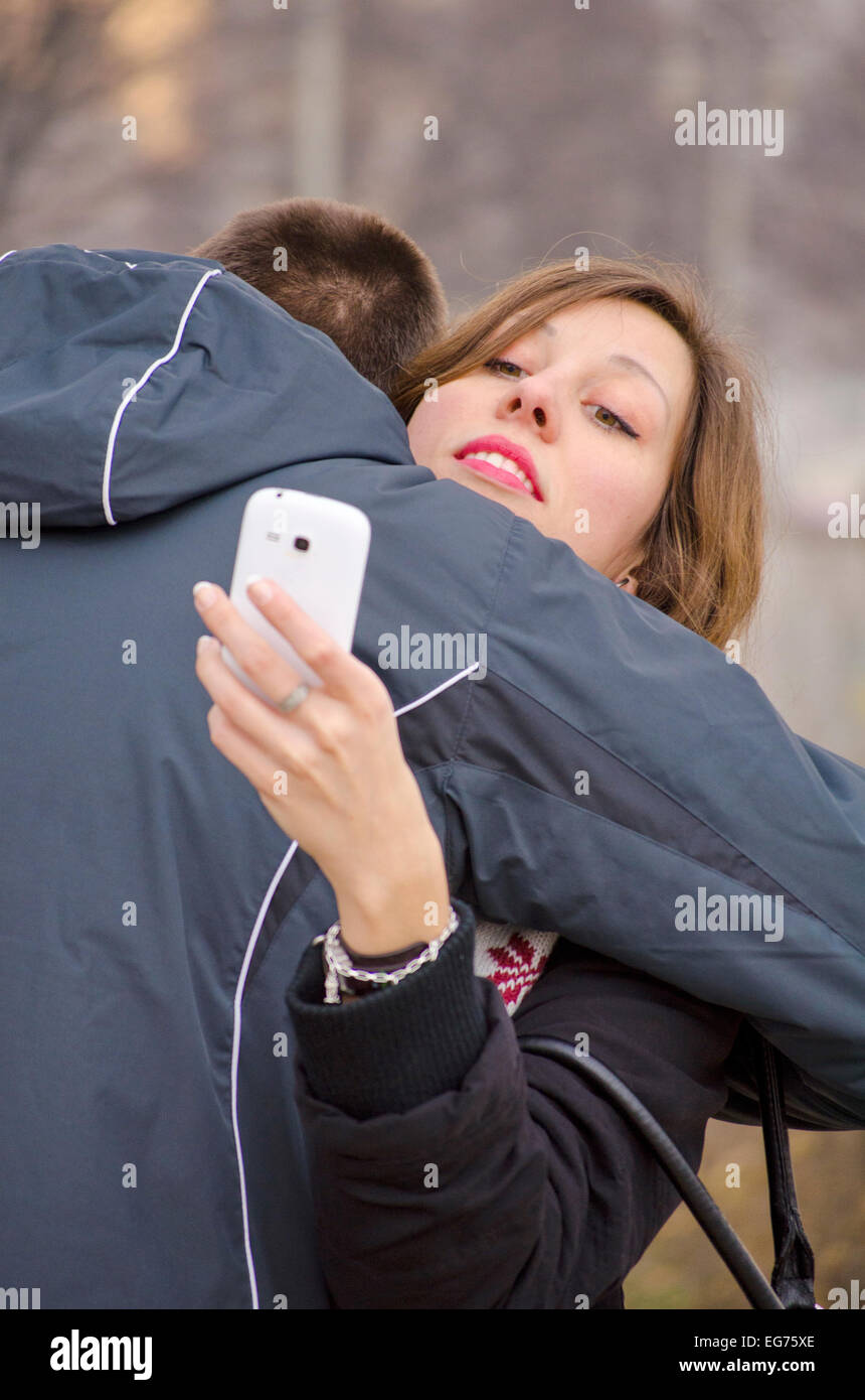 Junge, eine Mädchen umarmt, während sie ihr Smartphone im Freien sieht Stockfoto