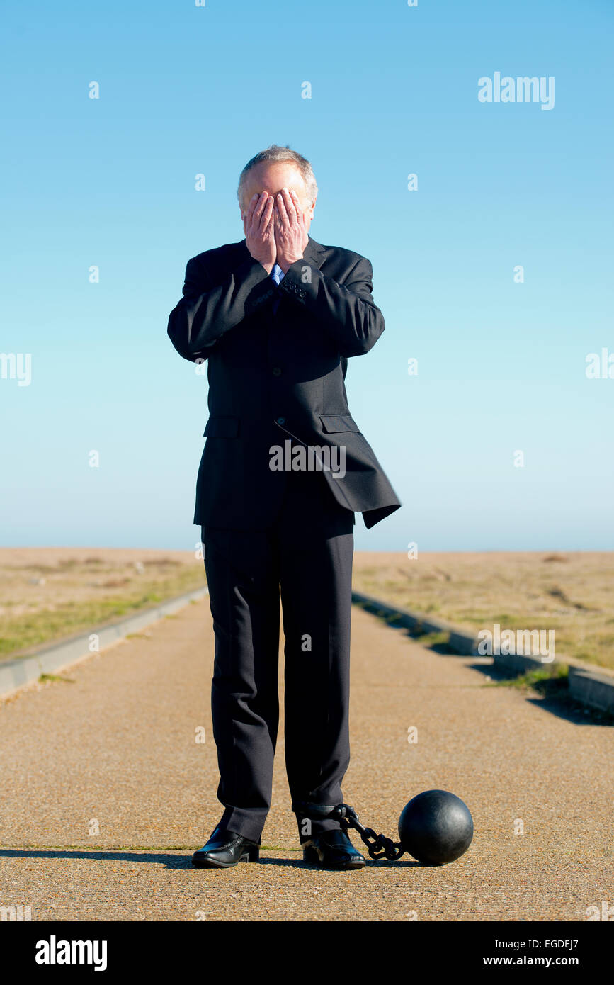 Kaufmann im dunklen Anzug, befestigt an einer Kugel & Kette mit seinem Kopf in seine Hände. Er steht in der Mitte einer abgelegenen Straße. Stockfoto