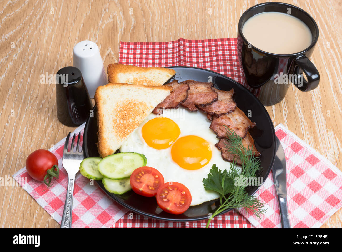 Spiegeleier mit Speck, Brot, Kaffee und Gemüse auf schwarzem Teller Stockfoto