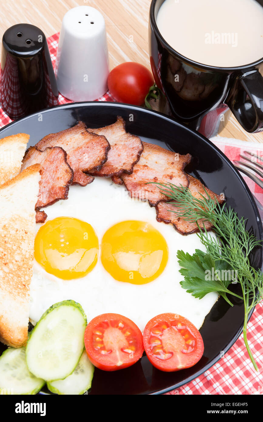 Spiegeleier mit Speck, Brot, Kaffee und Gemüse auf schwarzem Teller Stockfoto