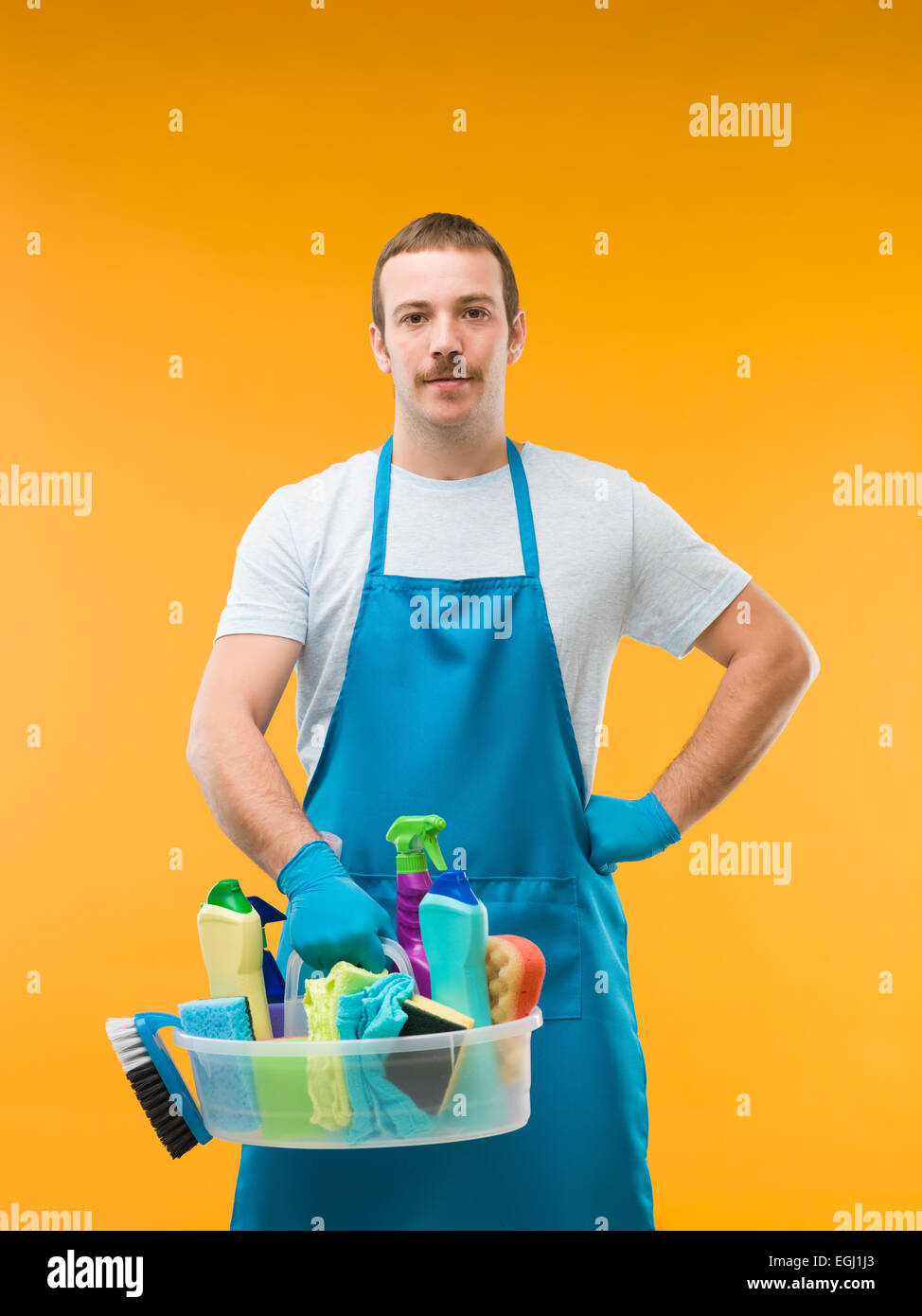 Kaukasische Hausmeister trägt Schürze und Holding-Korb mit Reinigungsmittel vor gelbem Hintergrund Stockfoto