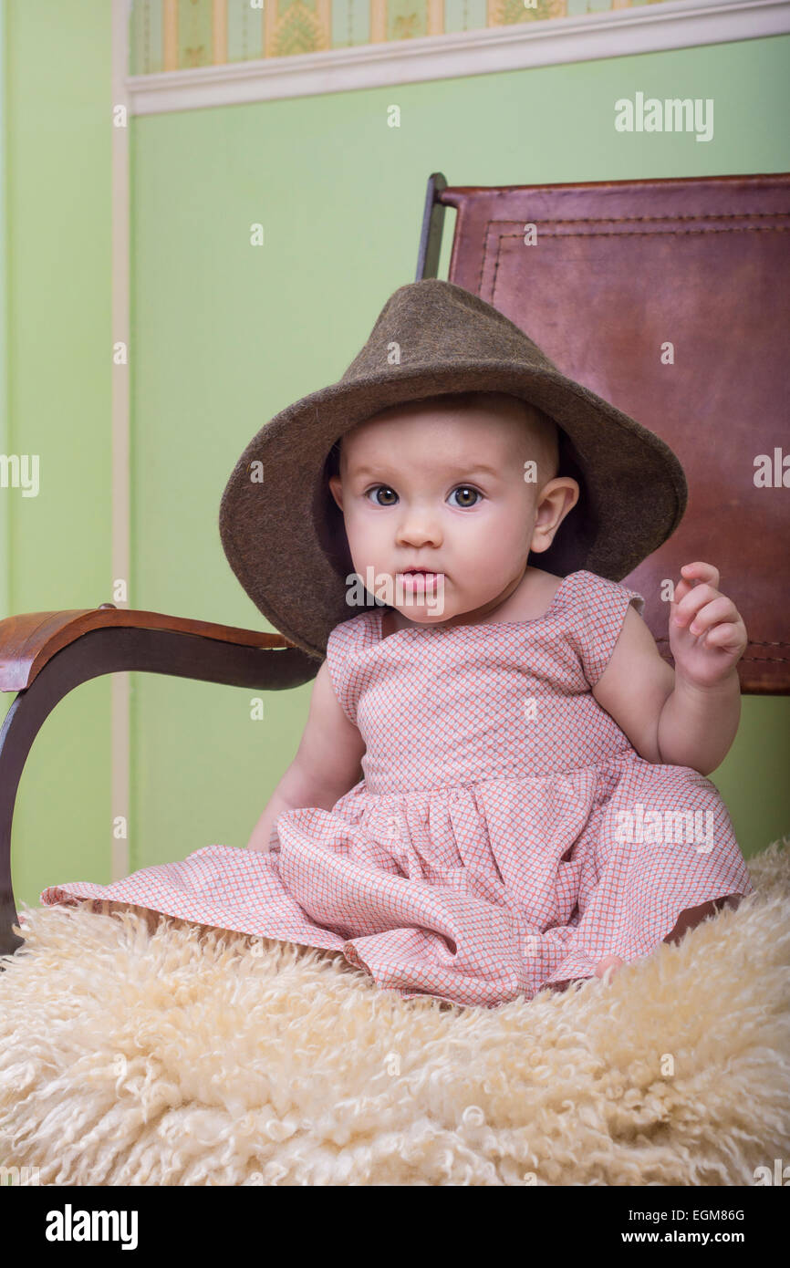 niedliche Baby tragen ein langes Kleid und Mutters Hut. Sitzend auf einem Stuhl im Zimmer Stockfoto