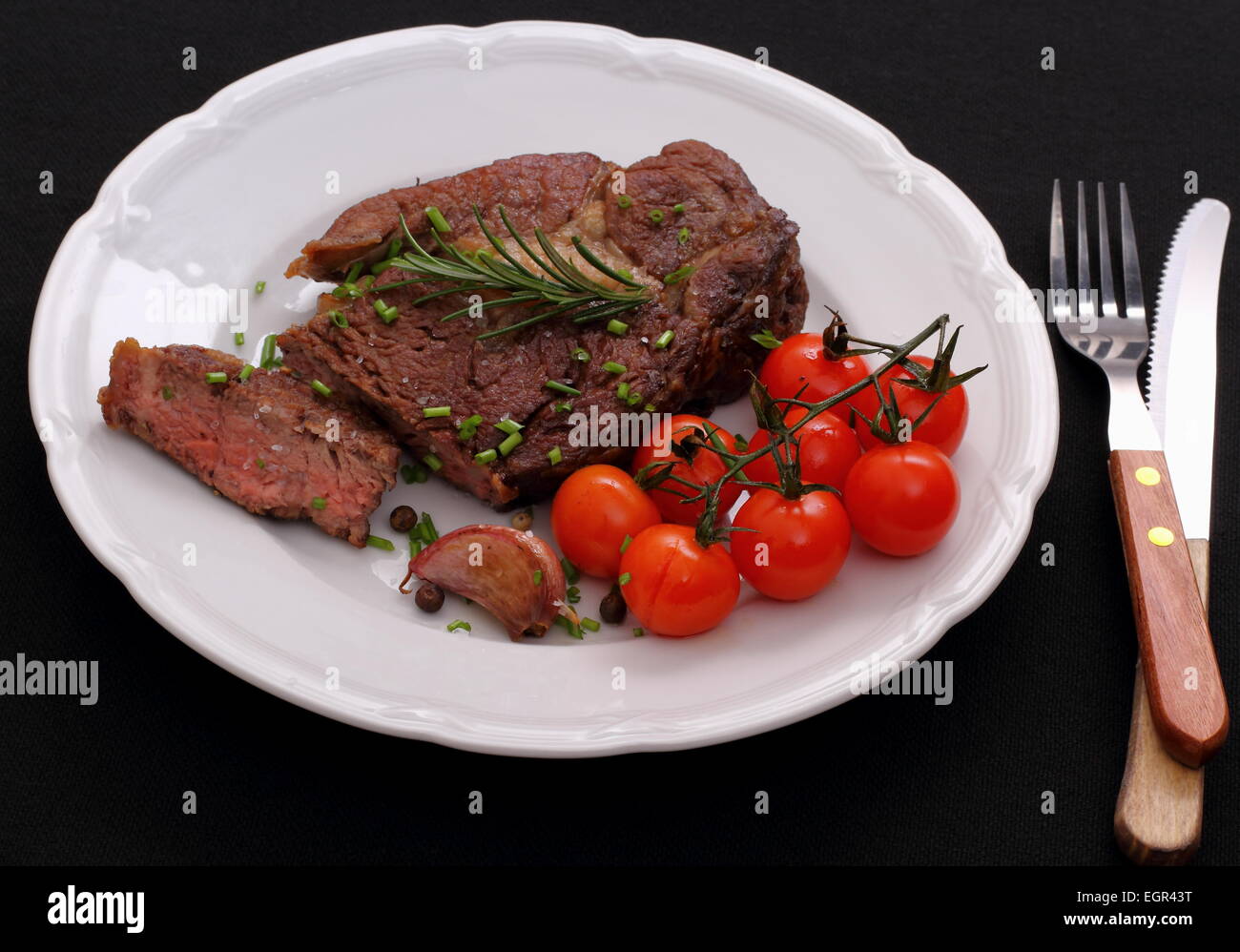 Rib-Eye Steak, Knoblauch, Kirschtomaten, Kräuter auf schwarzem Hintergrund, Ansicht von oben Stockfoto