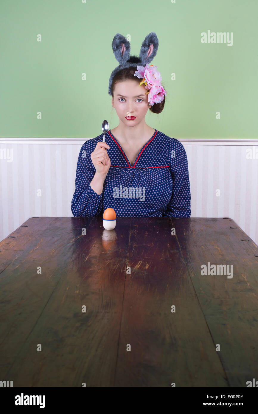 Porträt einer jungen Frau mit Hasenohren sitzt am Ende eine leere Tabelle mit Ei und Löffel. Stockfoto