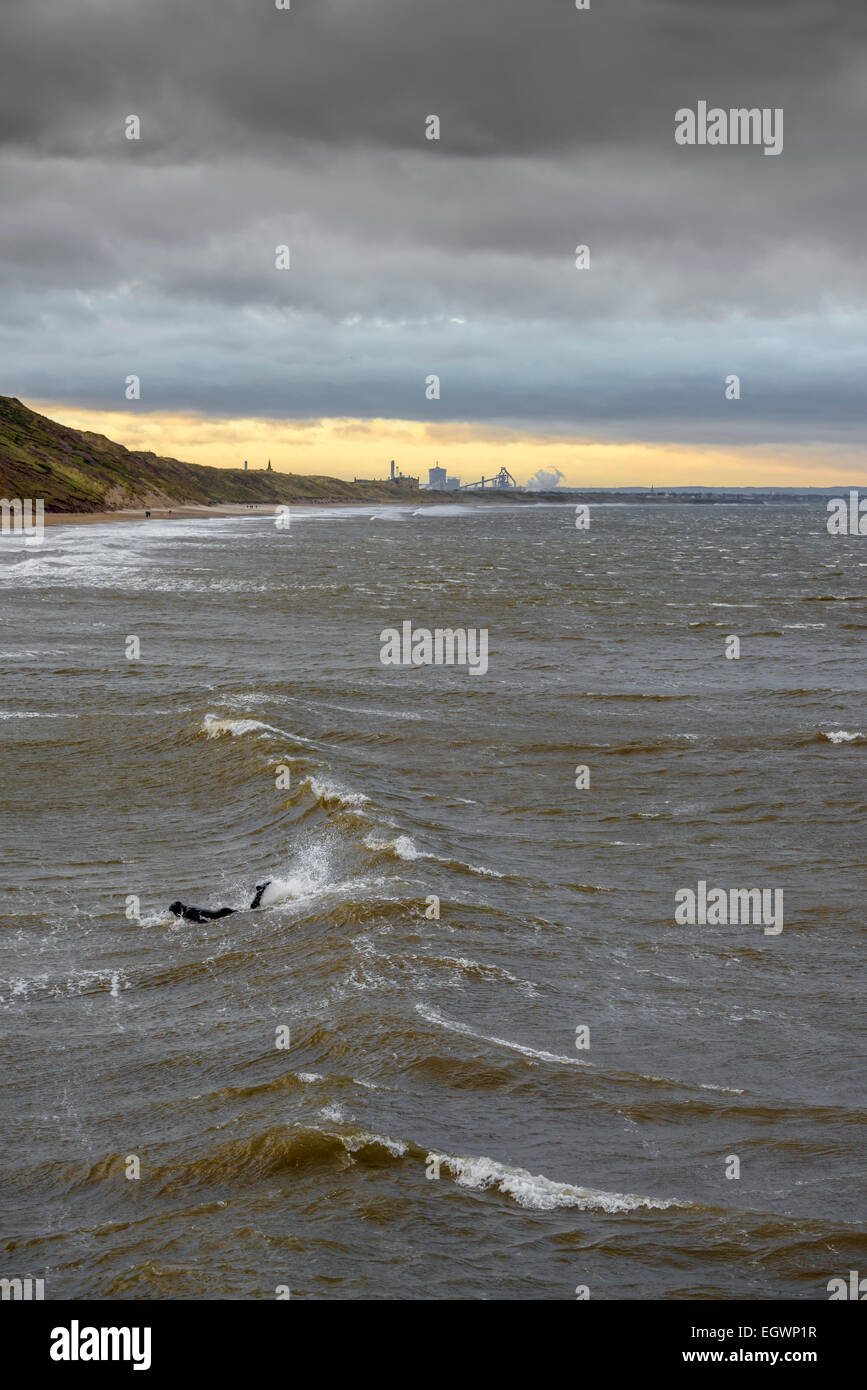 Einsamer Surfer Tritten in eine Welle an einem kalten Wintertag im Saltburn-by-the-Sea, North East UK mit einem Sonnenuntergang Himmel über Redcar fangen Stockfoto