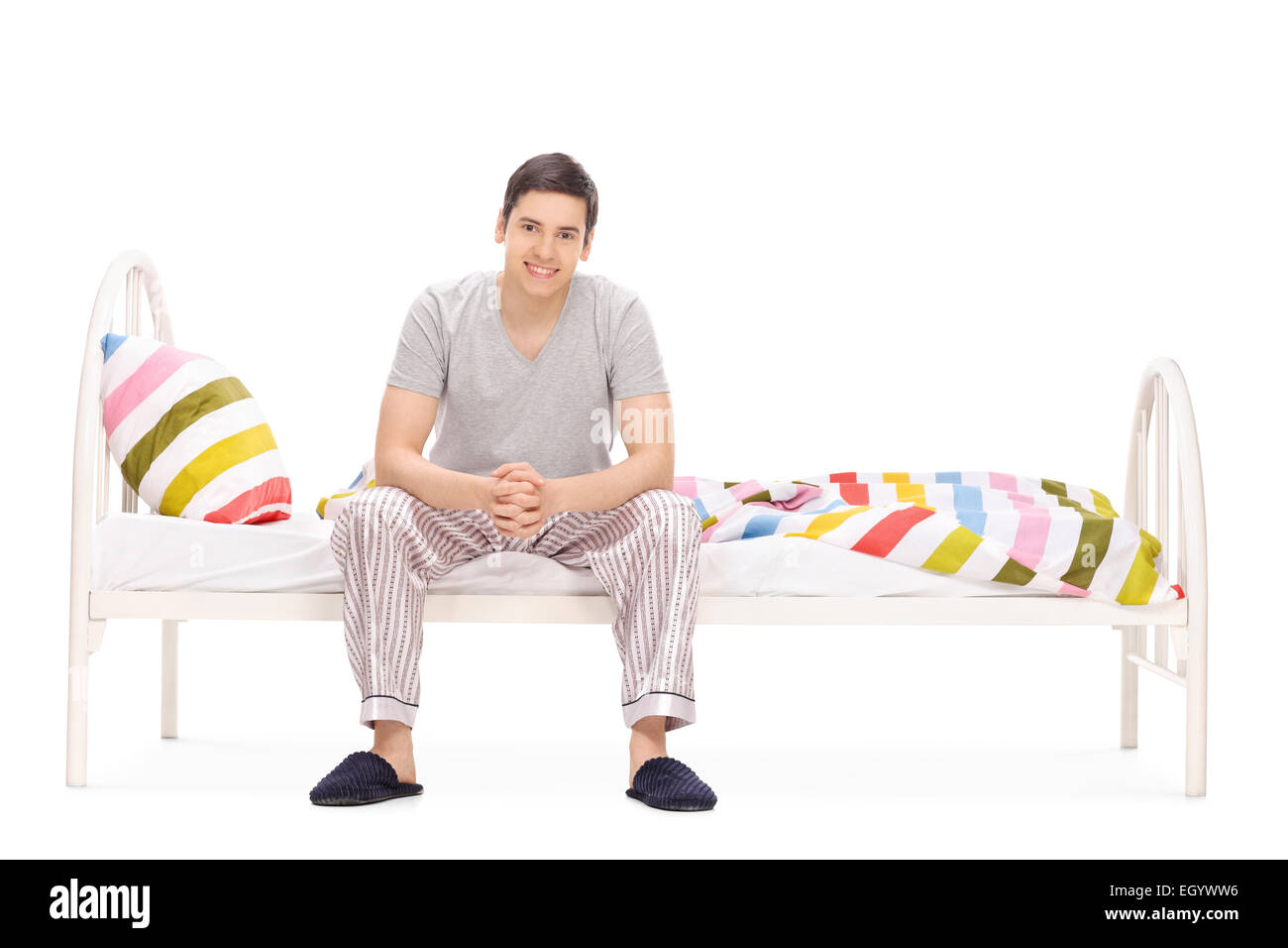 Fröhlicher junger Mann sitzt auf einem Bett isoliert auf weißem Hintergrund Stockfoto