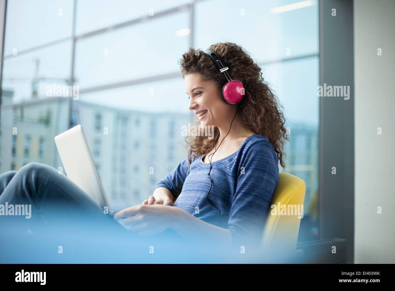 Lächelnde junge Frau tragen von Kopfhörern und mit Laptop am Fenster Stockfoto