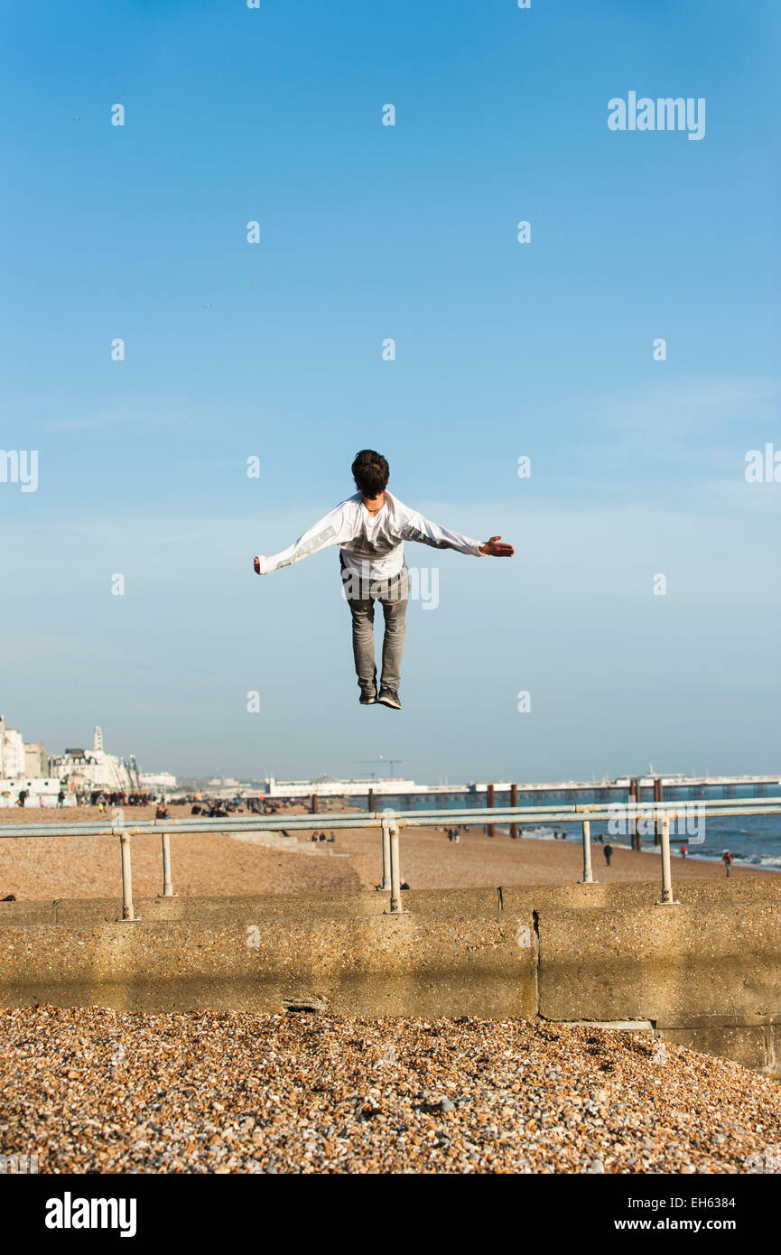 Ein junger Mann macht Purzelbäume und zurück kehrt in die Luft während eine Gruppe von jungen Menschen machen das Beste aus dem ersten Tag des sonnigen Frühlingswetter praktizieren die Kunst des Parkour, frei springen und Saltos, aus einem Steg auf Brighton Seafront mit Brighton Pier im Hintergrund zu tun. Brighton, East Sussex, England, UK. Stockfoto