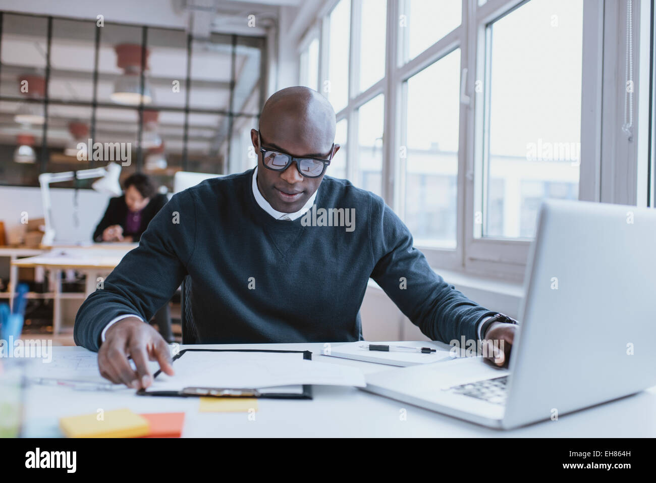 Junge afrikanische Führungskraft sitzt an seinem Schreibtisch mit Laptop Lesen eines Dokuments. Afrikaner, die im Büro arbeiten. Stockfoto