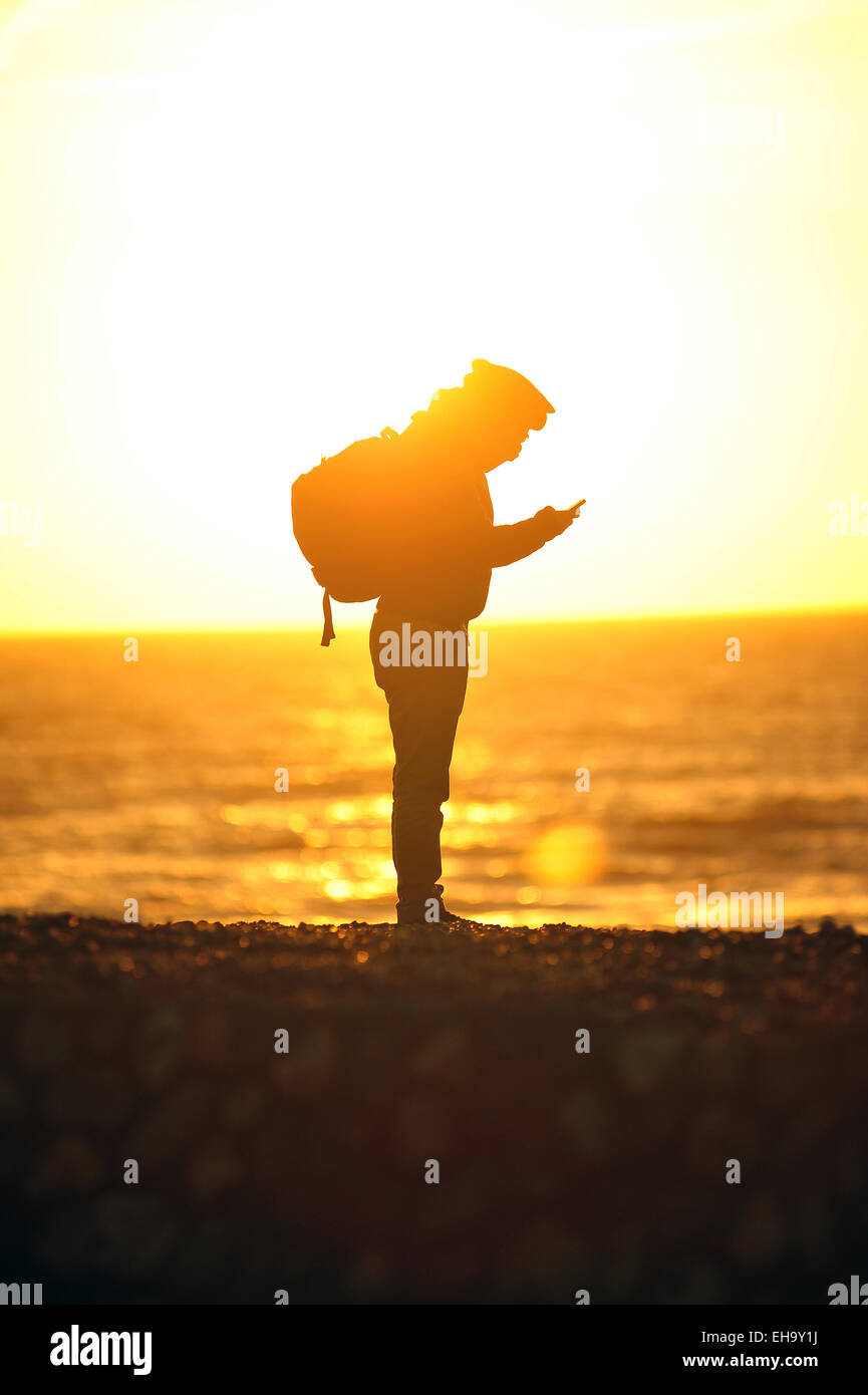 Eine Person ist gegen die Sonne Silhouette, wie sie ihr Telefon auf Brighton Beach betrachten. Stockfoto