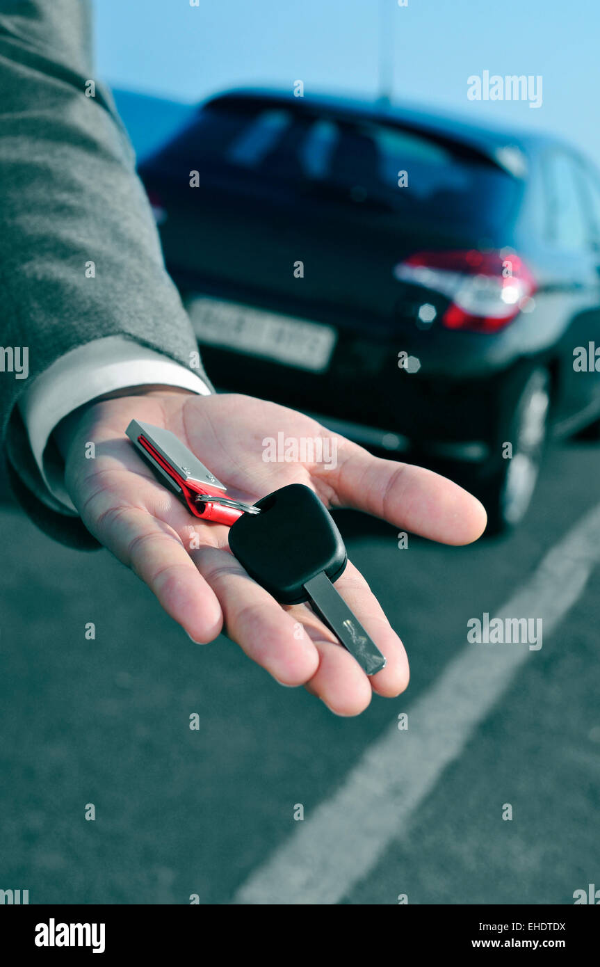 Mann im Anzug bietet einen Autoschlüssel für dem Betrachter mit einem Auto im Hintergrund Stockfoto