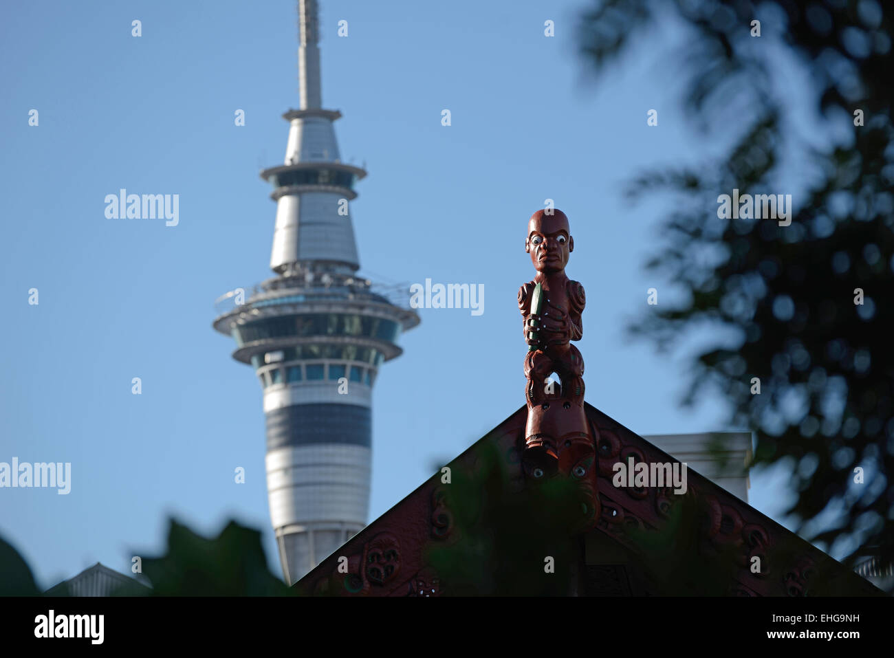 Schnitzwerks ein Maori Krieger steht Wache auf einem Marae, während die modernen Sky Tower die Skyline von Auckland in Neuseeland dominiert Stockfoto