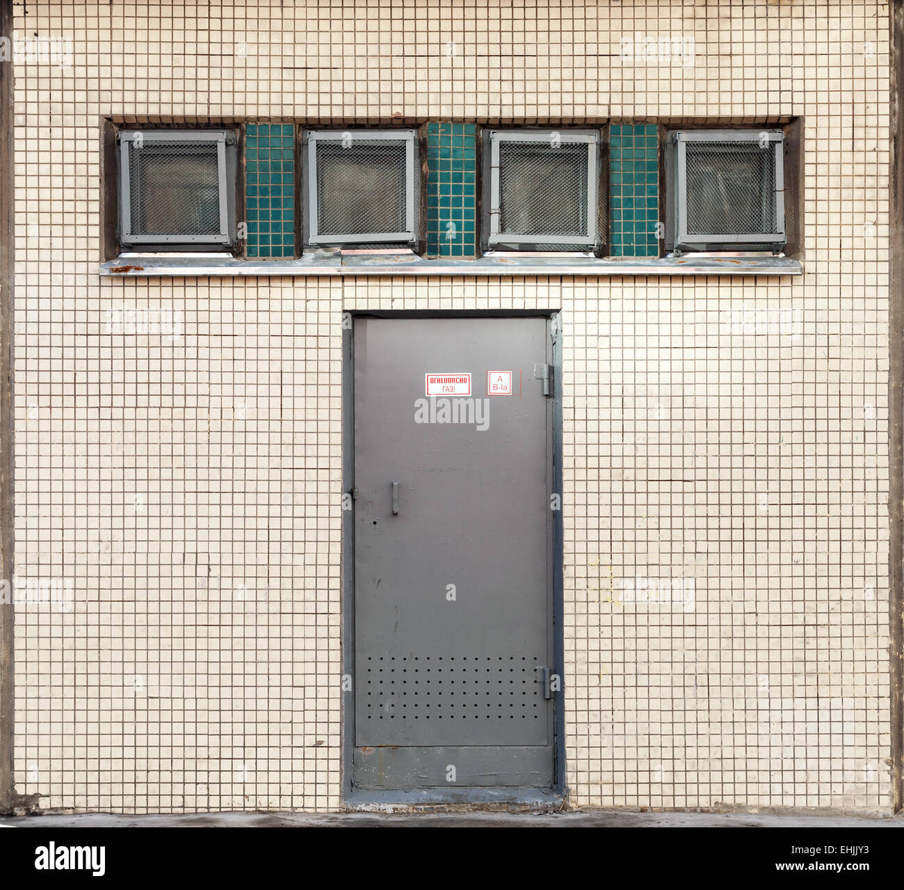 Wand Textur mit kleinen quadratischen Fenstern und Metalltür Industriegebäude. Russischen Text auf dem Etikett bedeutet brennbares gas Stockfoto