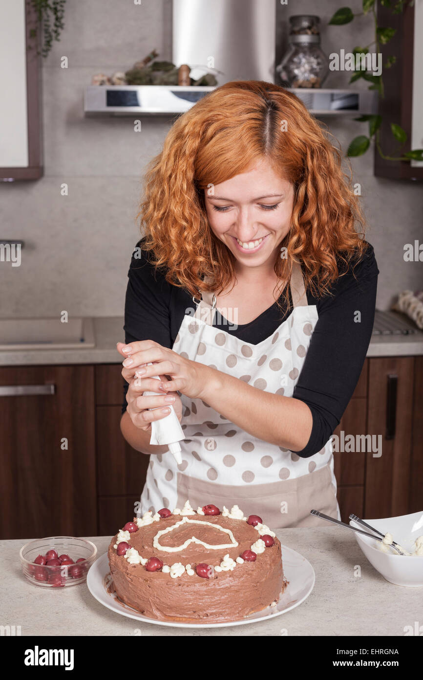 Glücklich rothaarige Frau Schokoladenkuchen mit Liebe Herzen, in der Küche zu Hause dekorieren. Stockfoto