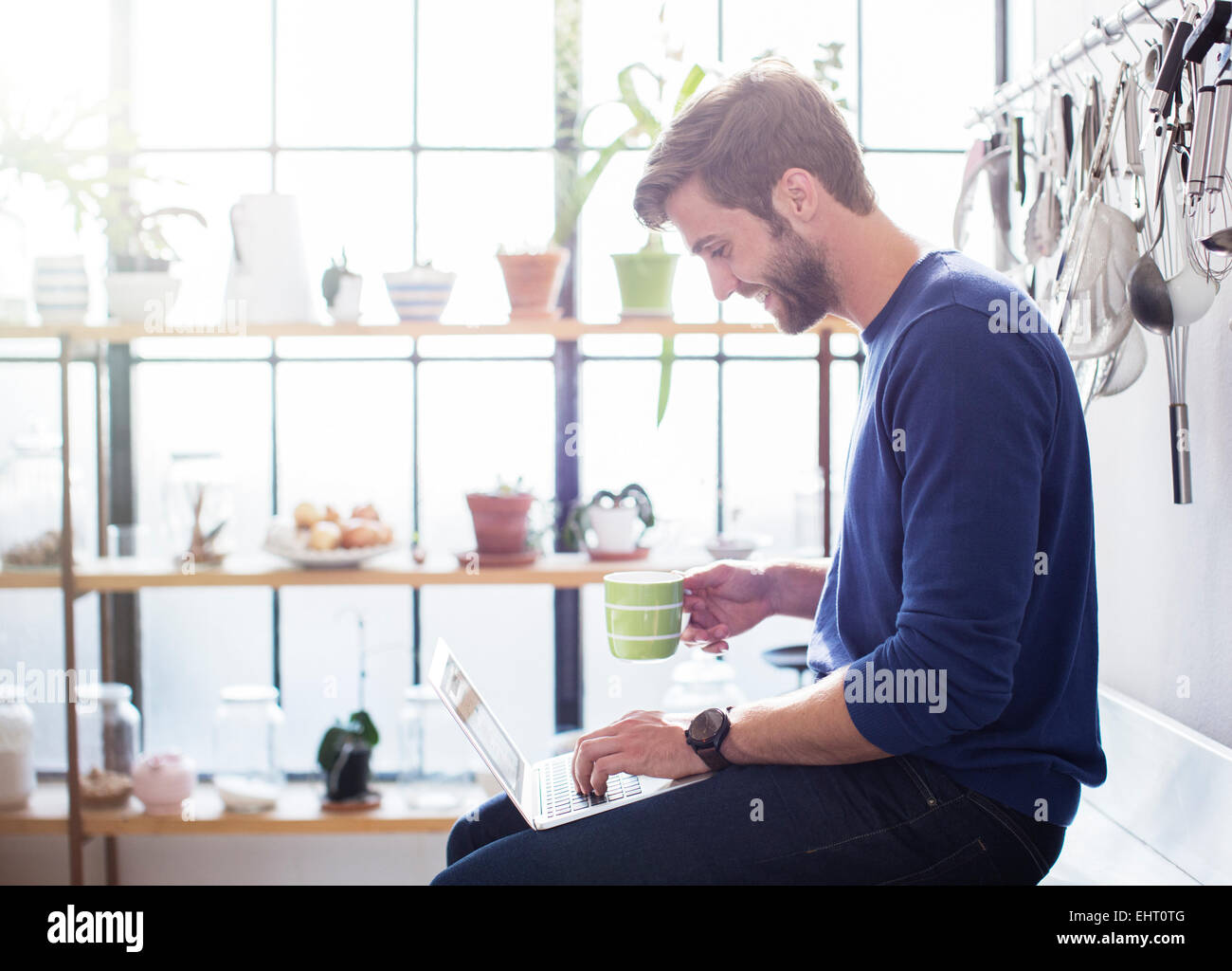 Junger Mann sitzt auf der Küchentheke mit Becher und laptop Stockfoto