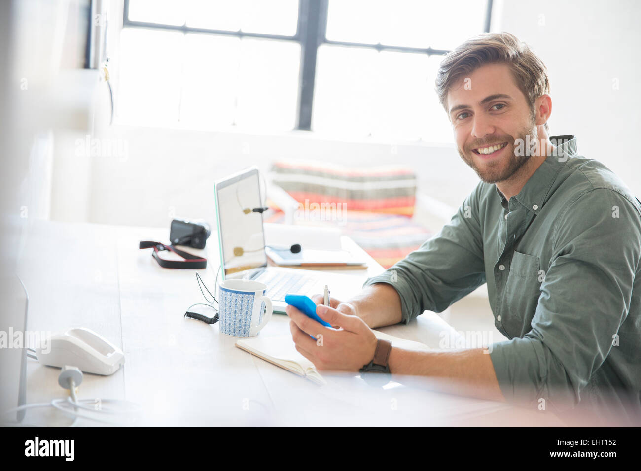 Porträt von junger Mann mit Handy und Laptop am Schreibtisch sitzen Stockfoto