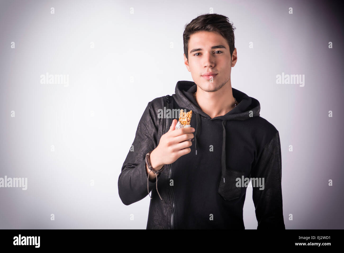 Hübscher junger Mann hält die Müsliriegel frisst er Kamera stehend, auf hellem Hintergrund zu betrachten, Stockfoto