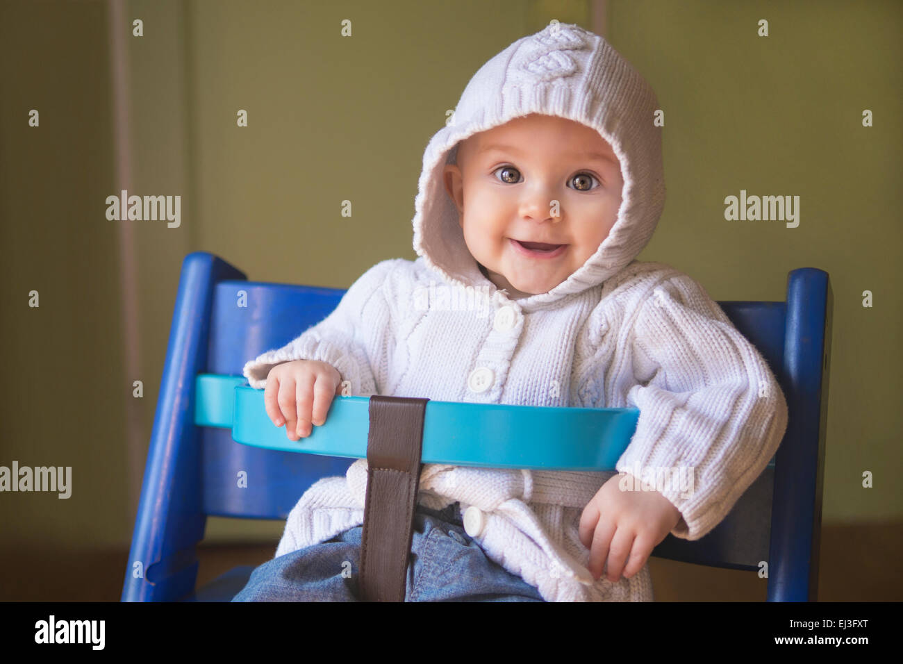 entzückende Babymädchen in einem weißen Kapuzenpullover sitzen in einem Kinderstuhl und lächelt in die Kamera. Stockfoto