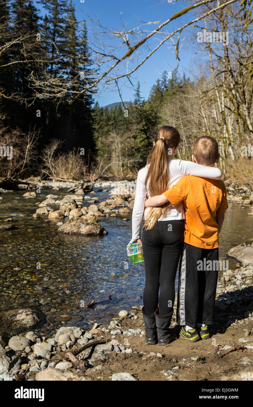 Neunjähriges Mädchen umarmt ihre sieben Jahre alten Bruder, wie sie innehalten und liebevoll, den Blick auf die flachen Snoqualmie River Stockfoto