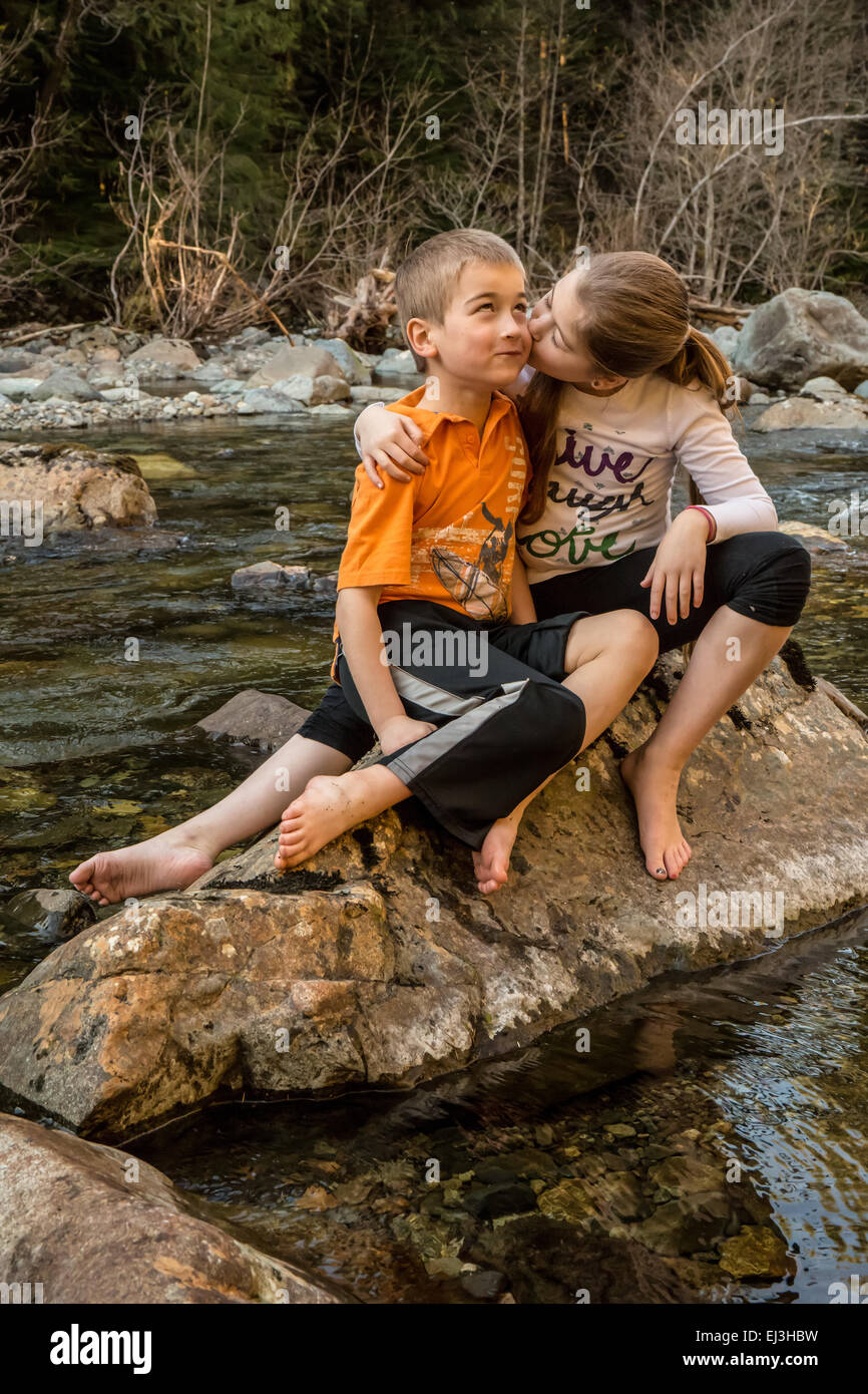 Neunjähriges Mädchen geben ihr sieben Jahre alten Bruder, eine Umarmung und einen Kuss beim Sitzen auf einem Felsen in einem seichten Fluss Stockfoto