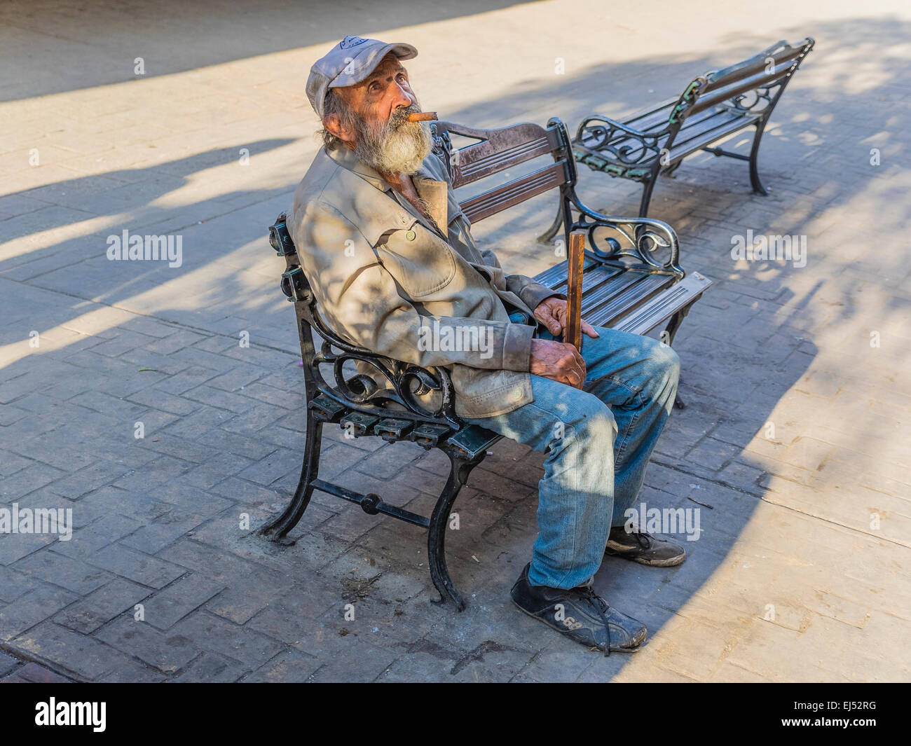 Eine kubanische männlichen Senioren mit einer Zigarre sitzt auf einer Bank mit einem Baseball-Typ-Hut, blickte in den Himmel im späten Nachmittag Licht. Stockfoto