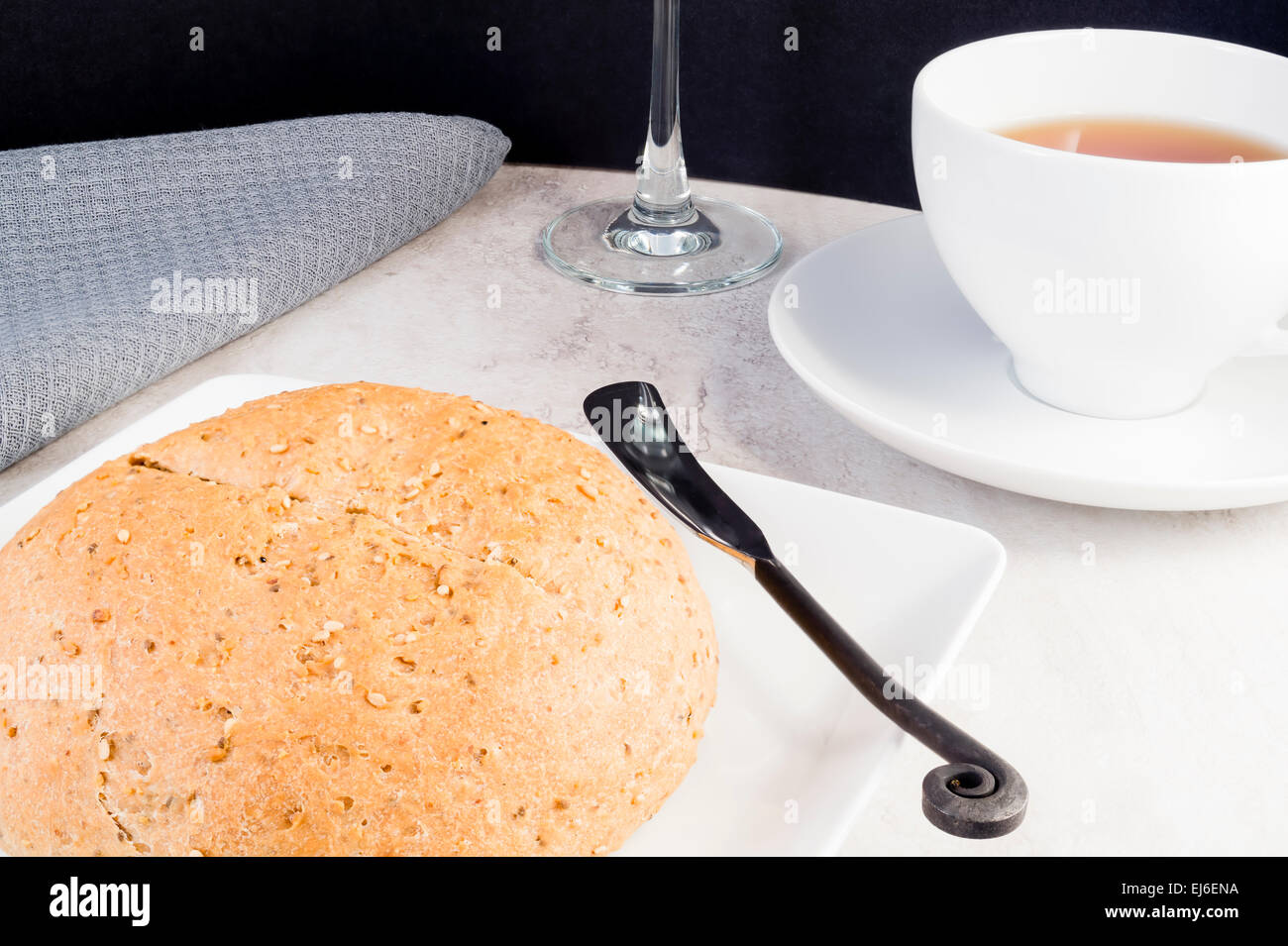 Rundes Laib Vollkornbrot mit Buttermesser, Tasse Tee und lange stemmed Glas auf Marmortisch mit grauen Serviette. Stockfoto