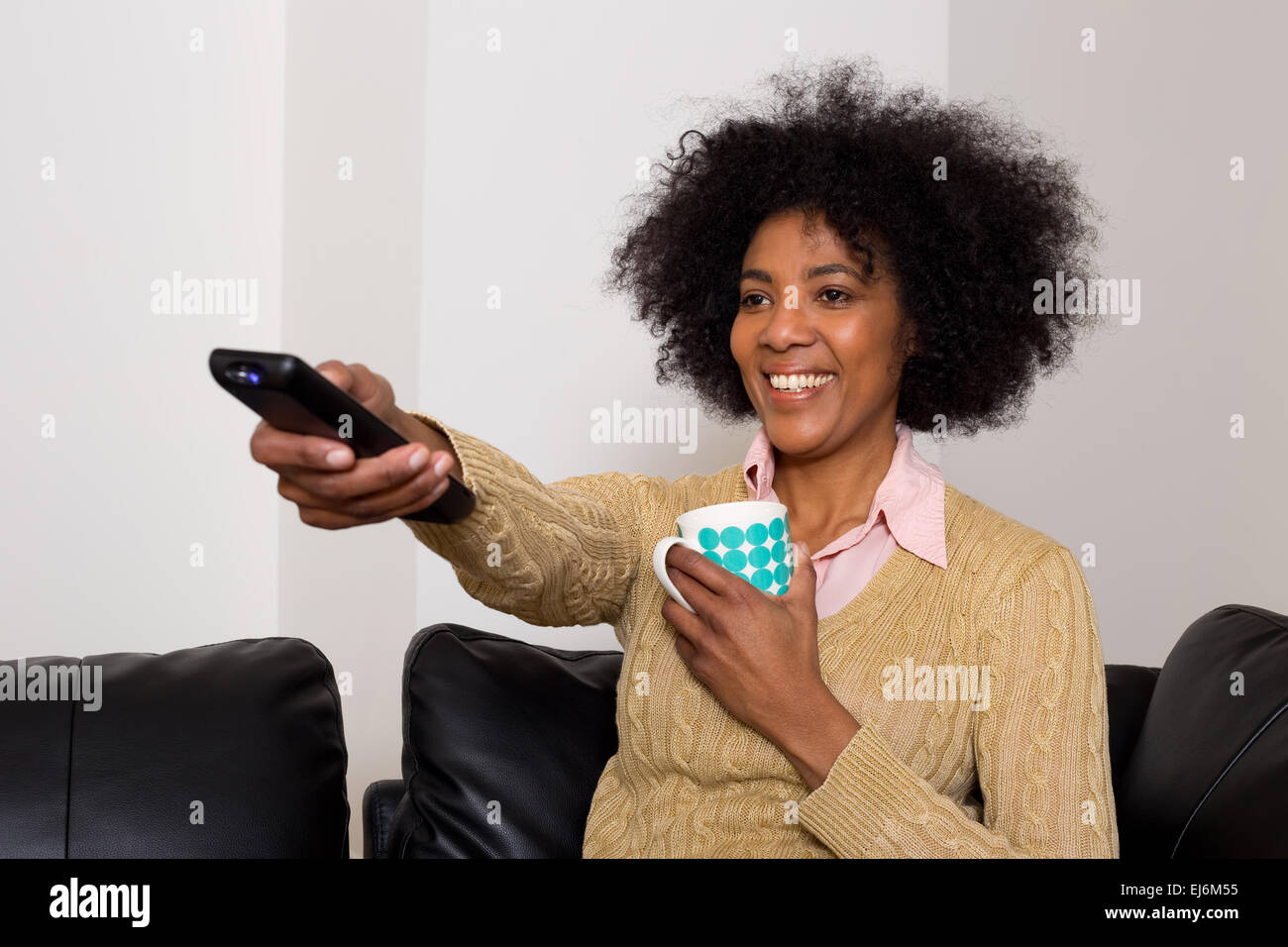 glückliche junge Frau genießen einen Kaffee und die Fernbedienung auf den Fernseher gerichtet. Stockfoto