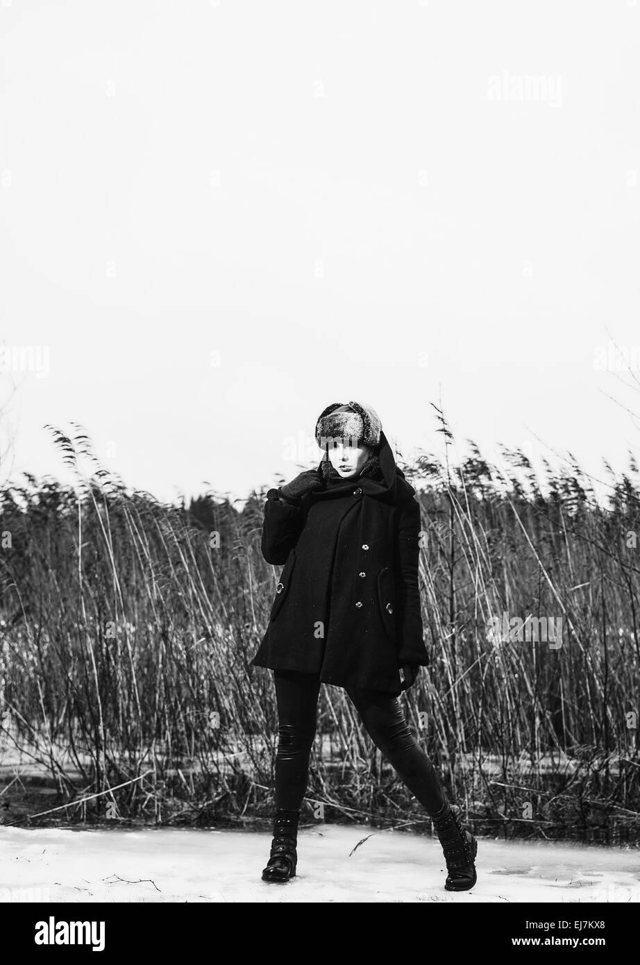 Mode Frau trägt einen Wintermantel und Pelzmütze und sie posiert vor dem Schilf, kalten regnerischen Wetters, schwarz in voller Länge und whi Stockfoto