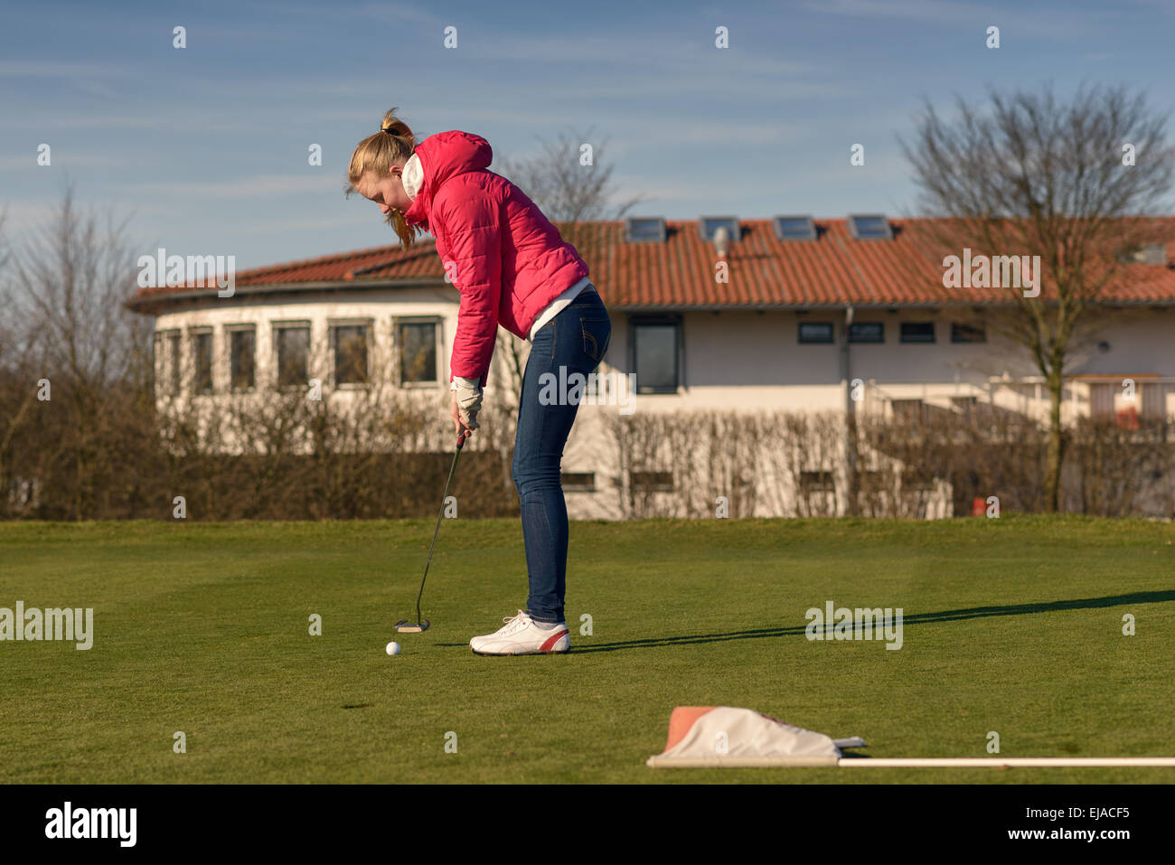 Junge weibliche Golfer tragen lässige Jacke und Jeans setzen Ball in Richtung des Lochs Stockfoto