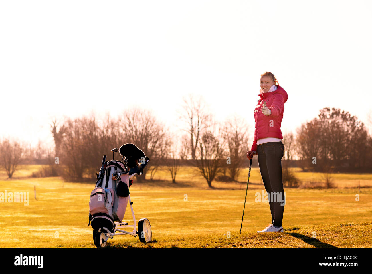 Junge weibliche Golfer in Jacke und Jeans ein Club, während ein Daumen mit Golf-Cart auf der Seite aufgeben. Stockfoto