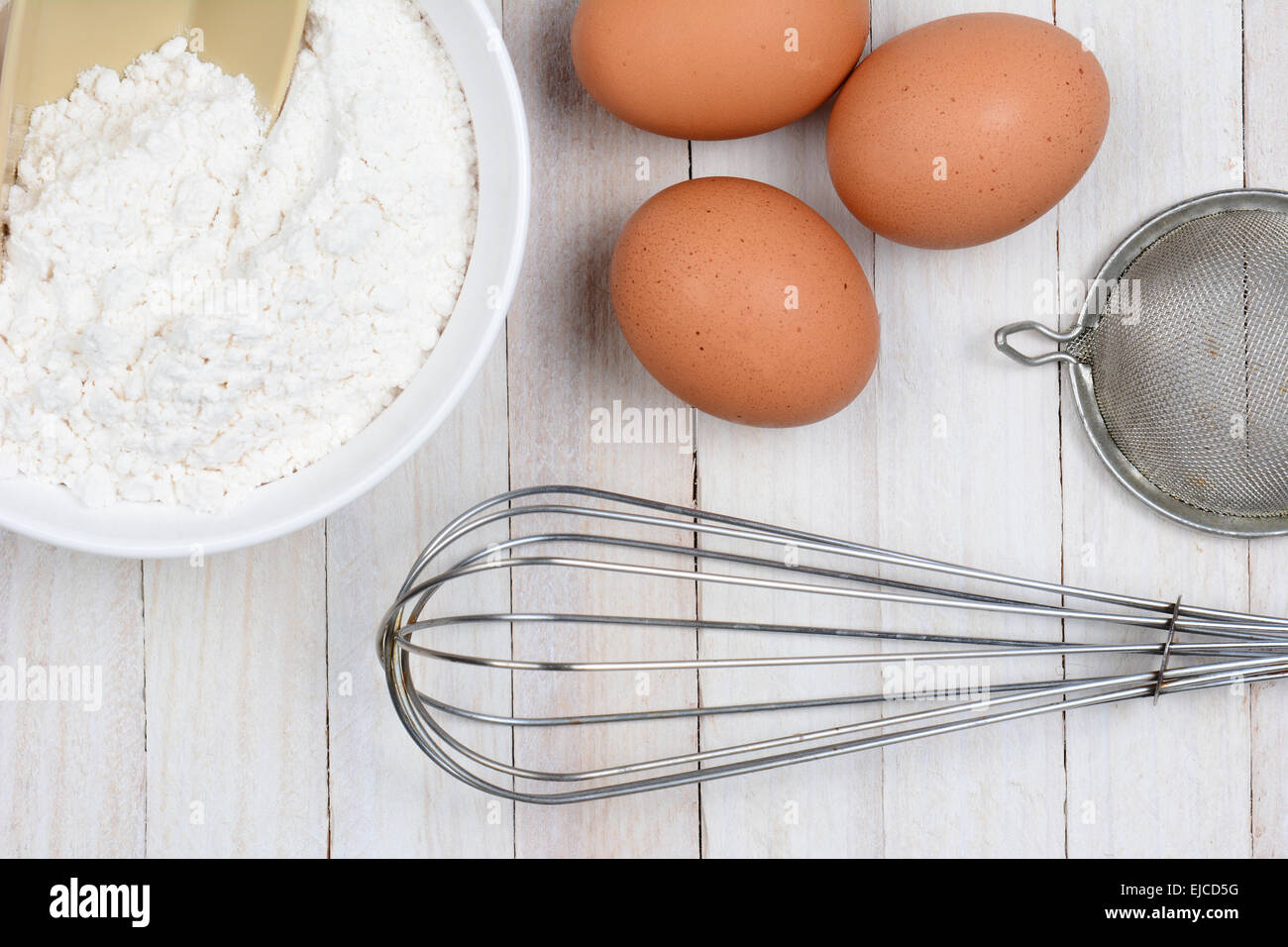 Hohen Winkel Closeup drei braune Eier, eine Schüssel mit Mehl, einen Schneebesen und Sieb auf einem rustikalen weißen Küchentisch. Stockfoto
