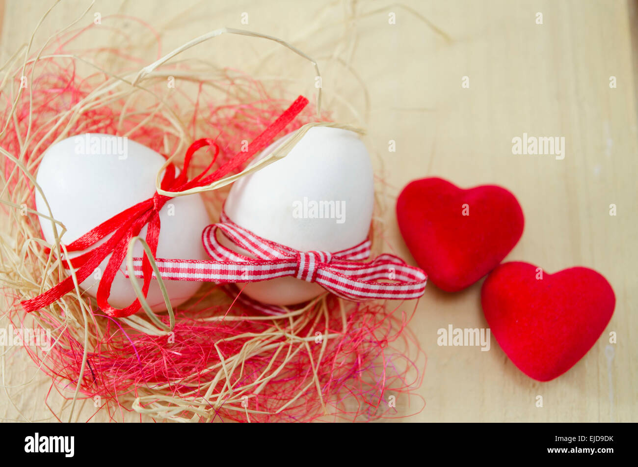 Die weißen Eiern gebunden mit roten Bändern in einem Nest liegend. Zwei kleine herzförmige Kissen auf einem Holztisch. Stockfoto
