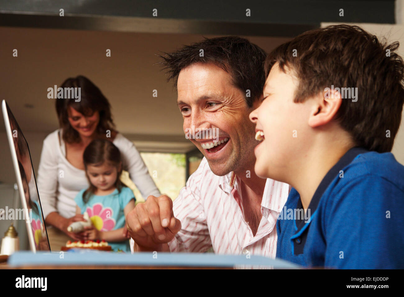 Eine Familie, zwei Eltern und zwei Kinder zusammen zu Hause. Vater und Sohn auf einem Laptop, und Mutter und Tochter einen Kuchen Glasur. Stockfoto