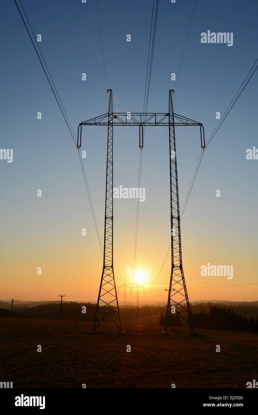 Hochspannungsleitungen mit großen und hohen Pylonen während des Sonnenuntergangs. Die Sonne geht zwischen zwei Pylonen. Stockfoto